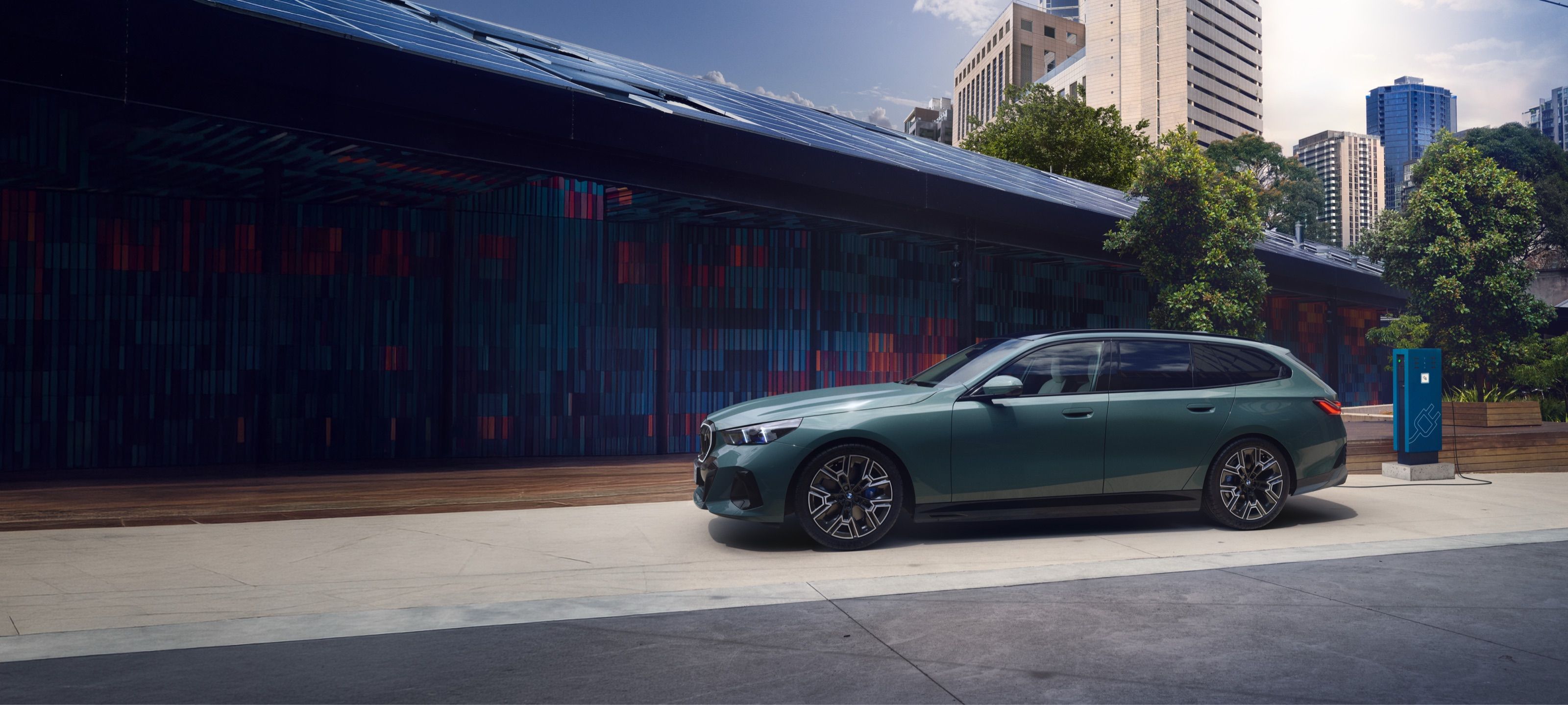 BMW i5 Touring in Grün an einer Ladestation vor einem modernen Gebäude