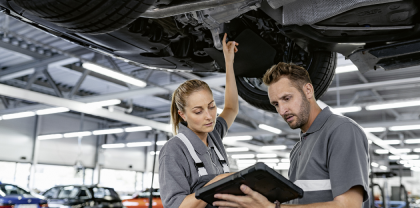 Ein weiblicher und männlicher Mechatroniker stehen unter einem Auto auf einer Hebebühne und schauen auf Tablet